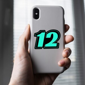 May include: A light gray phone case with the number "12" in a teal color with a black outline. The phone case is held in a person's hand.