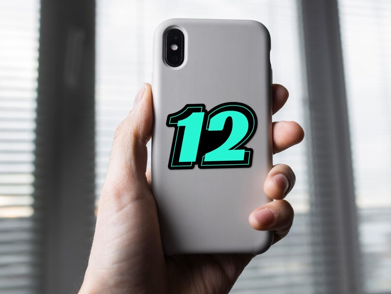 May include: A light gray phone case with the number "12" in a teal color with a black outline. The phone case is held in a person's hand.