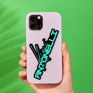May include: A light purple phone case with a black and teal graphic that reads "Antonelli". The graphic has a stylized design with lines and text. The phone case is held against a green background.