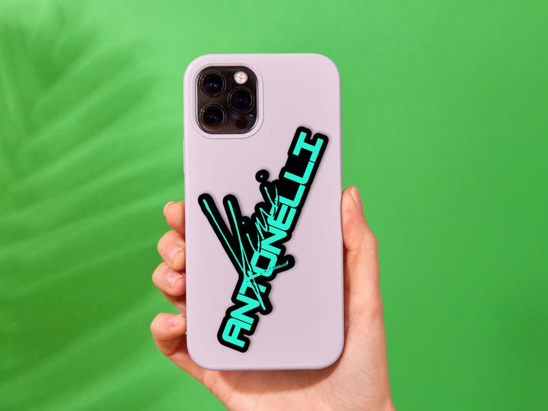 May include: A light purple phone case with a black and teal graphic that reads "Antonelli". The graphic has a stylized design with lines and text. The phone case is held against a green background.