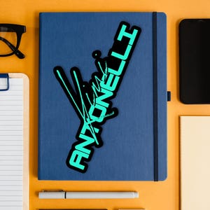May include: A blue notebook with a black and teal sticker that reads "Antonelli" in a stylized font. The notebook is surrounded by a black smartphone, glasses, a clipboard, a pen, and other stationery on an orange surface.