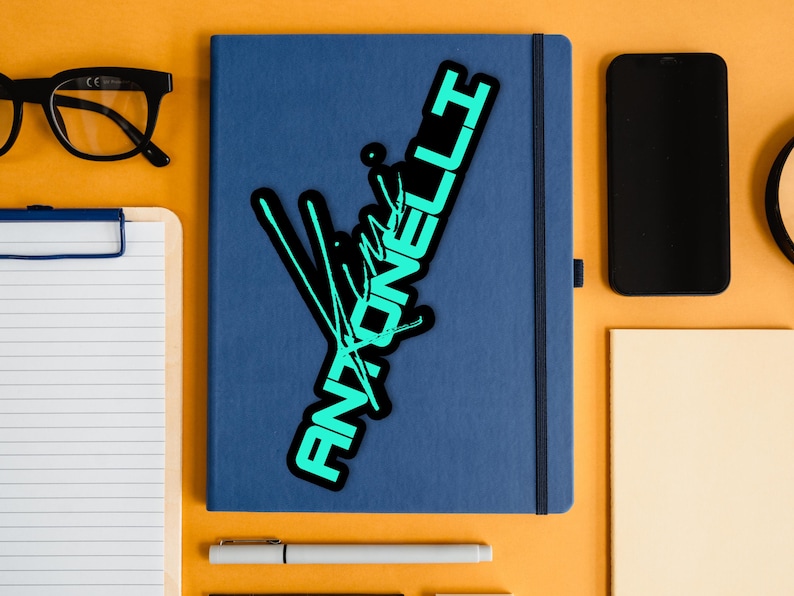 May include: A blue notebook with a black and teal sticker that reads "Antonelli" in a stylized font. The notebook is surrounded by a black smartphone, glasses, a clipboard, a pen, and other stationery on an orange surface.