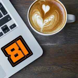 May include: A laptop computer with a sticker of the number 81 in orange and black on the lid. A cup of coffee with latte art is on a wooden surface next to the laptop.
