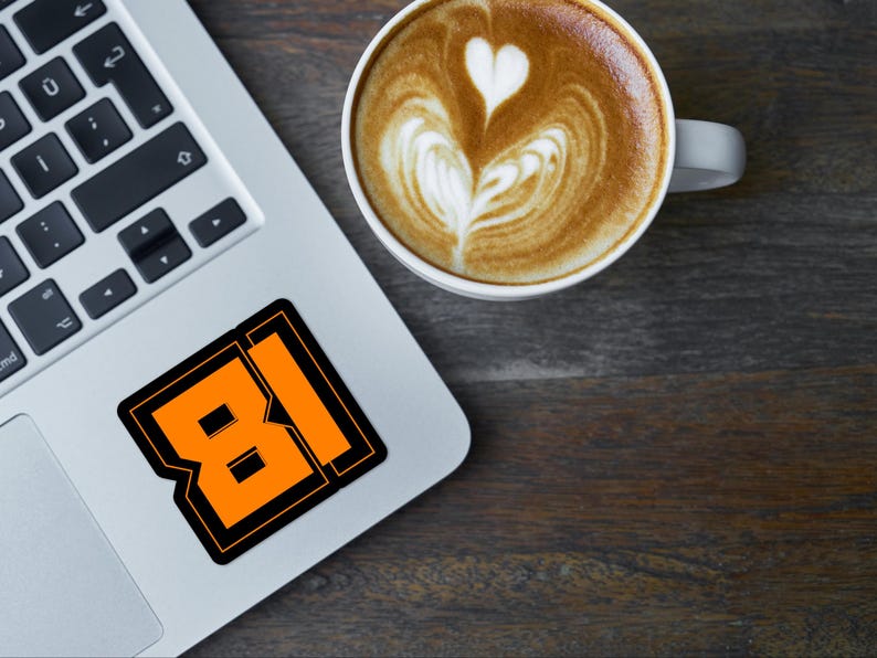 May include: A laptop computer with a sticker of the number 81 in orange and black on the lid. A cup of coffee with latte art is on a wooden surface next to the laptop.