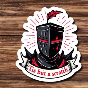 May include: A black and red knight helmet with a feather on top. The helmet has a red cross on the front and a white banner with the text "Tis but a scratch".