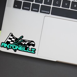 May include: A sticker featuring a checkered racing flag design with the name "Antonelli" in a teal font. The sticker is placed on a laptop, with a black background visible.