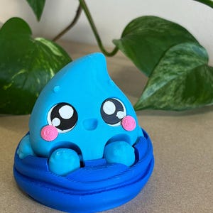以下が含まれることがあります： 大きな目とピンクの頬を持つ、青い漫画スタイルの水滴のフィギュアが、青い円形の容器の中に座っています。このフィギュアは、緑の葉を背景に配置されています。