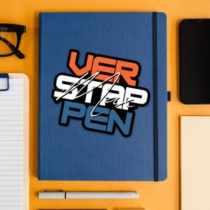 May include: A blue notebook with a sticker that says "VER STAP PEN" in white, orange, and blue lettering. The notebook is on a yellow surface with a pen, a notepad, and a smartphone.