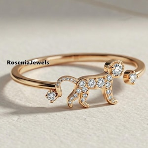 Könnte beinhalten: Ein goldfarbener Ring mit einem stilisierten Tierdesign, besetzt mit kleinen, klaren Edelsteinen. Der Ring hat ein zartes Band und einen Edelstein an jedem Ende der Tierfigur. Der Markenname "RoseniaJewels" ist sichtbar.