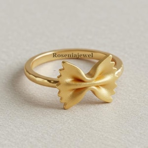 Pode incluir: Um anel dourado com um design de laço de massa farfalle. O anel tem uma banda lisa e uma forma de laço detalhada. O nome da marca "Roseniajewel" está impresso no anel.