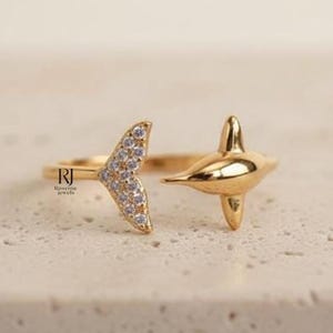 May include: A gold-colored ring featuring a dolphin design. The ring has a dolphin tail with small, clear stones and a dolphin body. The ring is open, allowing for an adjustable fit. The ring is on a light-colored surface.