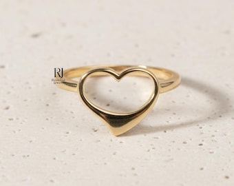 May include: A gold-colored ring with a heart-shaped design. The ring features a hollow heart shape, with a smooth, polished finish. The ring is set against a neutral background, highlighting its simple elegance.