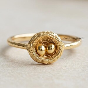 Op de afbeelding: Een goudkleurige ring met een nestontwerp met twee kleine, ronde gouden bollen in het midden. De ringband heeft een textuurpatroon en het nest heeft een geweven uiterlijk. De ring staat tegen een zachte, neutrale achtergrond.