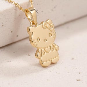 Peut inclure: Un collier doré avec un pendentif en forme de chat de dessin animé avec un nœud. Le pendentif présente des détails de traits du visage et de vêtements. La chaîne est délicate et dorée, créant un accessoire charmant.