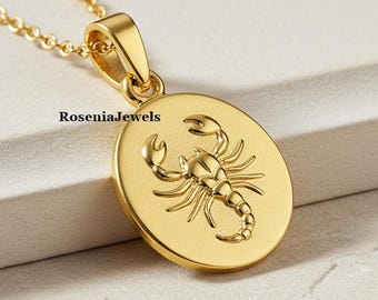 May include: A gold-colored pendant necklace featuring a detailed scorpion design within an oval frame. The pendant hangs from a delicate chain. The brand name "RoseniaJewels" is printed on the pendant.