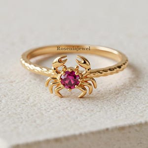 Pode incluir: Um anel dourado com um design de caranguejo e uma pedra preciosa vermelha redonda no centro. A banda do anel tem um padrão texturizado. O nome da marca "Rosentajewel" está gravado no anel.
