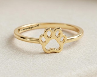 Puede incluir: Un anillo dorado con un diseño de huella de pata. El anillo tiene una banda delgada y una huella de pata recortada en el centro. El texto "Roseniajewel" está grabado en la banda. El anillo está sobre un fondo neutro.