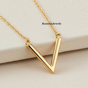Könnte beinhalten: Eine goldfarbene V-förmige Anhängerkette. Der Anhänger hat eine solide V-Form und hängt an einer filigranen Kette. Die Halskette wird auf einer hellen Oberfläche präsentiert. Der Text "RoseniaJewels" ist in der oberen rechten Ecke sichtbar.