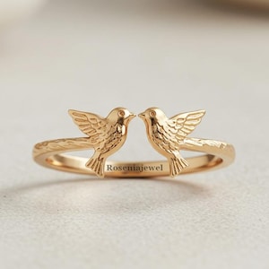 Könnte beinhalten: Ein goldfarbener Ring mit zwei detaillierten Vogelfiguren, die sich zugewandt sind. Der Ring hat ein strukturiertes Band und der Markenname "Roseniajewel" ist auf der Vorderseite eingraviert. Die Vögel haben ausgebreitete Flügel und sind so positioniert, als würden sie sich gleich mit den Schnäbeln berühren.