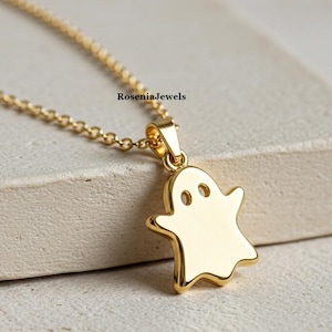 Könnte beinhalten: Eine goldfarbene Geister-Anhänger-Halskette. Der geisterförmige Anhänger hat zwei Augenlöcher und eine glatte Oberfläche. Die Halskette ist ebenfalls goldfarben. Der Markenname "RoseniaJewels" ist oben rechts sichtbar.