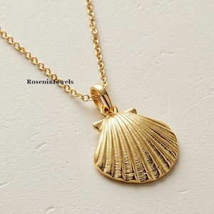 Puede incluir: Un collar con colgante de concha dorada. El colgante presenta un diseño de concha detallado y texturizado. La cadena del collar es delicada y dorada. El fondo es de color blanco roto neutro. El nombre de la marca "Rosenia Jewels" es visible.
