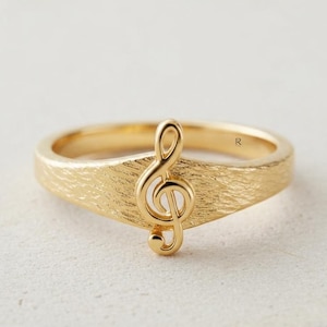 May include: A gold ring featuring a textured band and a prominent treble clef symbol centered on the front. The metal surface has a brushed, matte finish, and the musical note design is raised, creating a distinct focal point on the band.