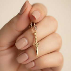 May include: A gold-colored pendant necklace featuring a detailed dagger design. The dagger has an ornate hilt and a pointed blade. The necklace is held by a hand with manicured nails. The jewelry is from "Rosonia Jewels".