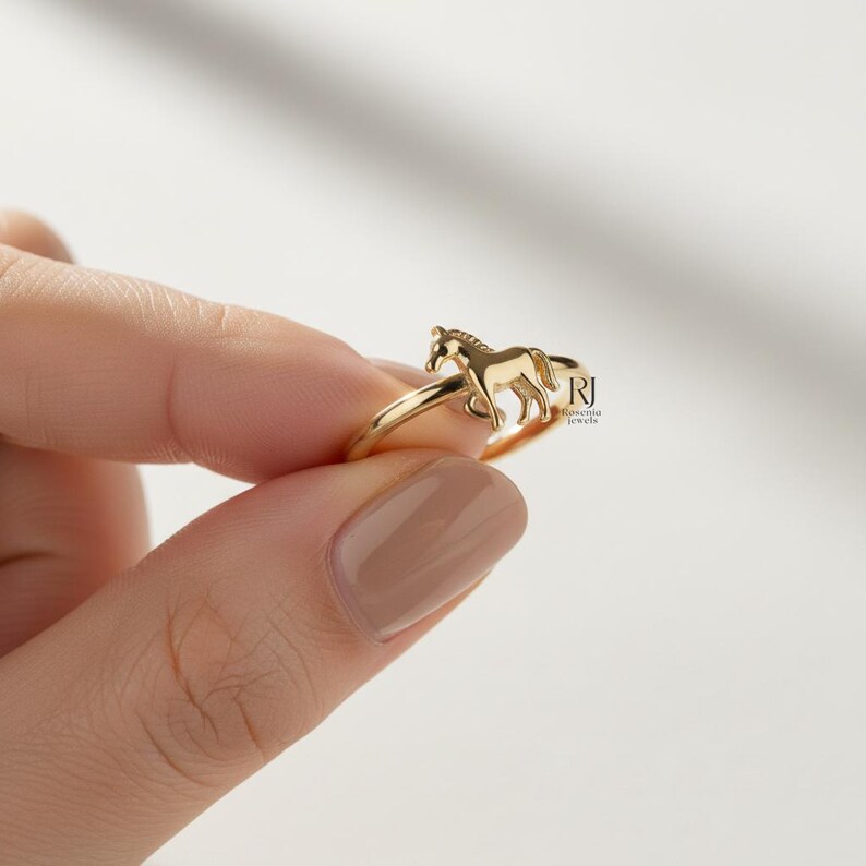 May include: A gold-colored ring featuring a detailed horse design. The ring is held by a hand against a white background. The horse is depicted in a running pose, adding a dynamic element to the jewelry.
