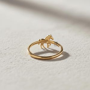 May include: A gold-colored ring featuring a detailed horse figure. The horse is positioned on the ring's band, with its legs and body visible. The ring has a polished finish and is set against a neutral background.