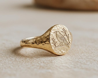 May include: Gold signet ring with a hammered band and an oval face. The face features an engraved raven perched on a branch with bare tree branches. The ring is a warm gold color.