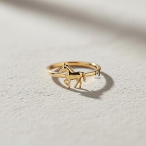 May include: A gold-colored ring featuring a horse-shaped charm. The ring has a thin band and a detailed horse figure in a walking pose. The ring is set against a neutral background, highlighting the jewelry's design.