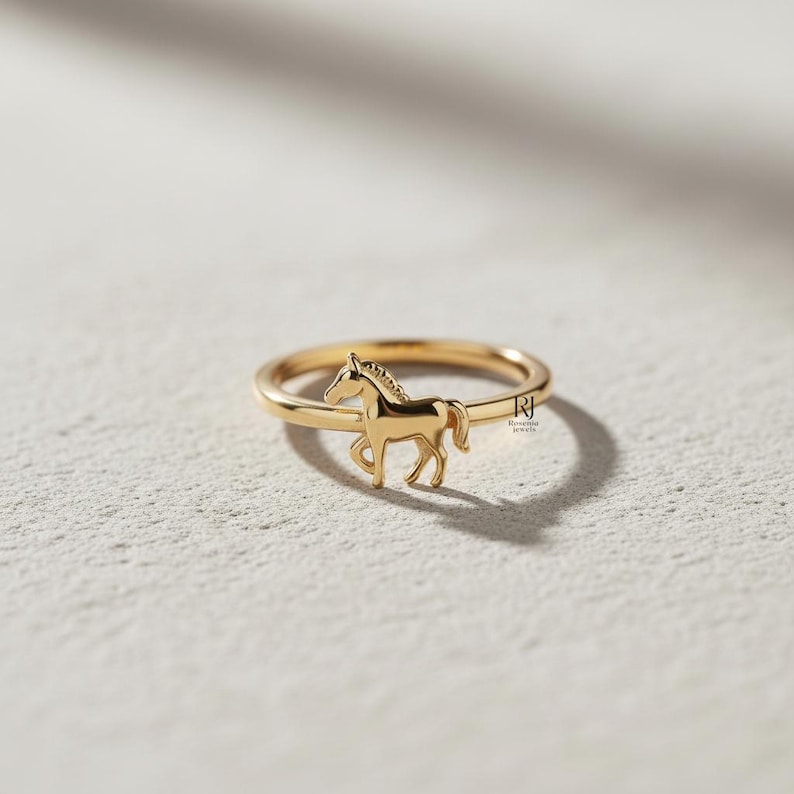 May include: A gold-colored ring featuring a detailed horse silhouette. The ring has a thin band and a small, three-dimensional horse figure. The horse is depicted in a running pose, adding a touch of elegance to the jewelry.