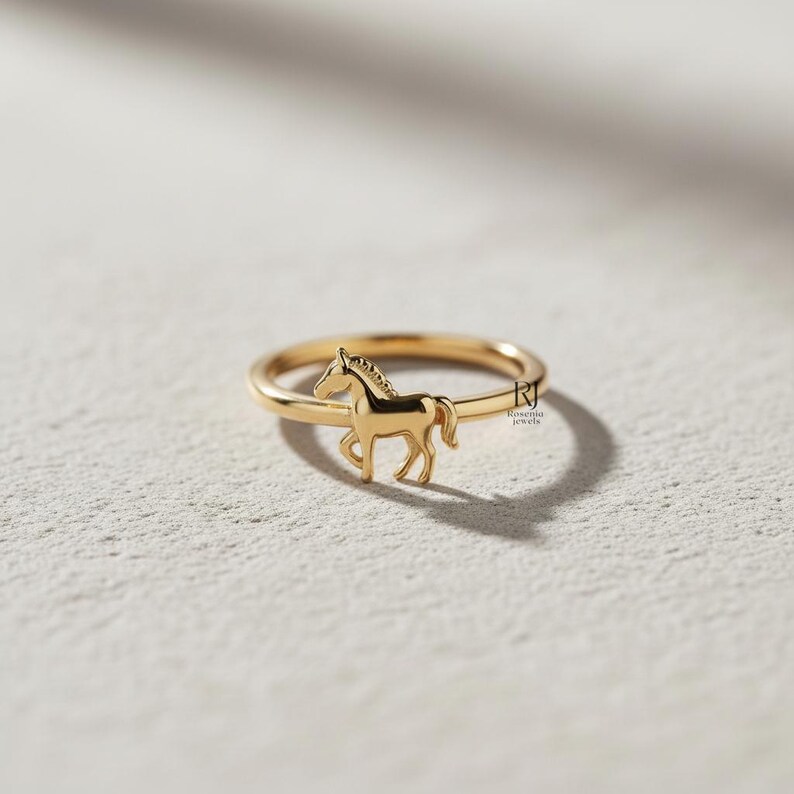 May include: A gold-colored ring featuring a horse silhouette. The ring has a thin band and a detailed horse figure in a walking pose. The ring is set against a neutral background, highlighting the jewelry's design and craftsmanship.