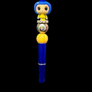 May include: A blue pen with a decorative design featuring a cartoon character figure at the top. The pen has a series of colorful beads, including a yellow sphere and a marbled bead. The pen is set against a black background.