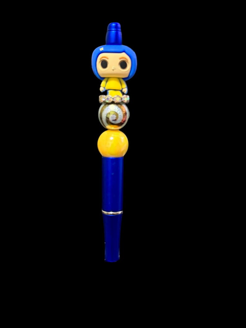 May include: A blue pen with a decorative design featuring a cartoon character figure at the top. The pen has a series of colorful beads, including a yellow sphere and a marbled bead. The pen is set against a black background.