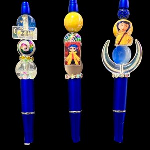 May include: Three blue pens with decorative tops. One pen has a clear top with the word "Coraline" and a rainbow bead. Another features a yellow bead and a character image. The third has a blue bead and a crescent moon design.