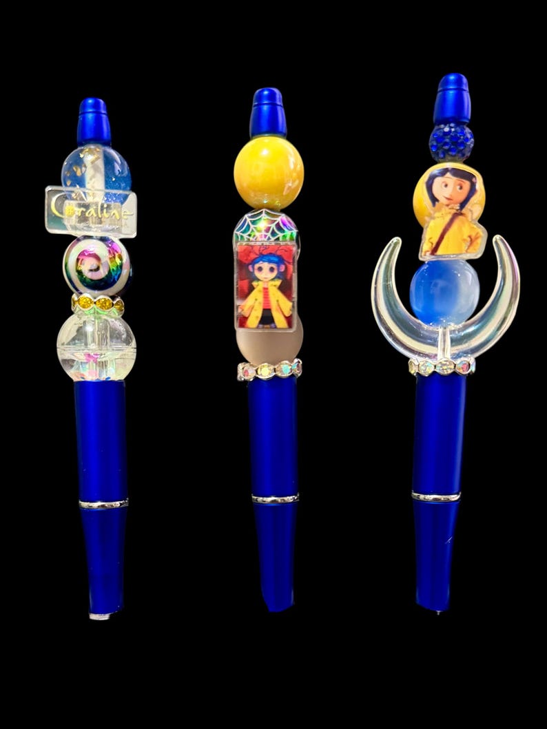May include: Three blue pens with decorative tops. One pen has a clear top with the word "Coraline" and a rainbow bead. Another features a yellow bead and a character image. The third has a blue bead and a crescent moon design.