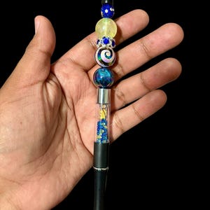 May include: A black pen with a decorative beaded top. The beads are blue, yellow, and iridescent, with a swirl design. The pen barrel contains blue and yellow beads and a small figure.