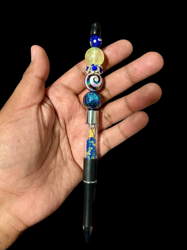 May include: A black pen with a decorative beaded top. The beads are blue, yellow, and iridescent, with a swirl design. The pen barrel contains blue and yellow beads and a small figure.