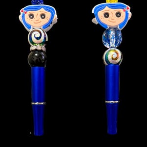May include: Two blue pens with decorative tops featuring a cartoon character with blue hair and a white face. The pens have a series of colorful beads and a silver band. The background is black.