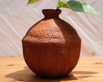 May include: A terracotta vase with a rough, textured surface. The vase has a rounded shape and a dark brown rim.