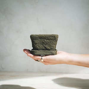 May include: A hand holding a small, dark green, textured ceramic planter with a matching saucer.