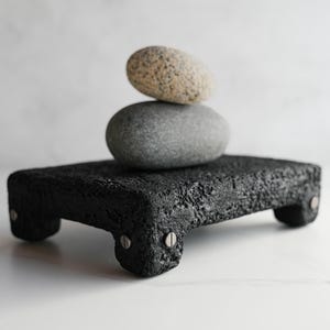May include: A rectangular black volcanic rock platform with four short legs attached by silver metal screws. Two smooth, rounded river stones are stacked on top of the platform, with a speckled beige stone resting on a larger, solid grey stone.