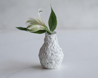 May include: A white ceramic vase with a textured surface, holding a single white flower with green leaves.