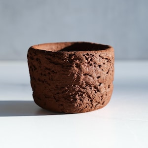 May include: A small, cylindrical, handmade terracotta pot with a rough, textured surface. The dark brown ceramic vessel is unglazed and has a natural, earthy appearance. The pot is empty and sits on a white surface.