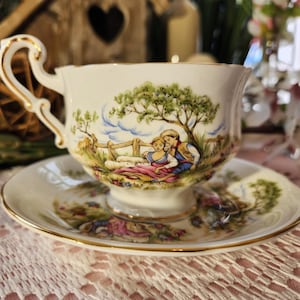 May include: A white teacup and saucer set with a gold trim. The cup and saucer feature a pastoral scene with a couple under a tree. The handle of the cup has a decorative gold design.