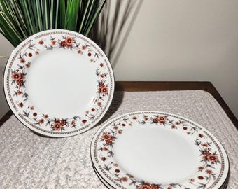 May include: Two white ceramic dessert plates with a floral pattern in shades of brown and red. The plates have a gold trim around the edge.