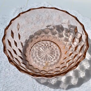 May include: A vintage pink glass bowl with a diamond-cut pattern and a scalloped rim. The bowl has a circular design in the center and is likely used for serving or display. The bowl is made of glass and is a light pink color.