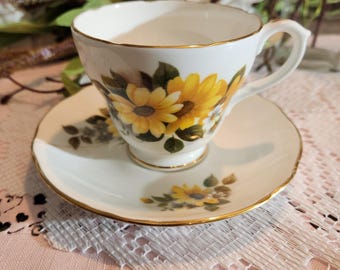 May include: A white teacup and saucer set with a gold rim. The teacup and saucer are decorated with yellow sunflowers and green leaves. The teacup is sitting on a white saucer with a matching floral design.