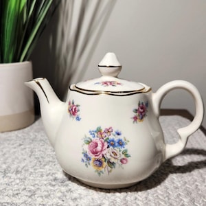 May include: A white ceramic teapot with a floral design featuring pink, blue, and yellow flowers. The teapot has a gold trim and a curved handle. The lid is topped with a small finial. A decorative item for tea parties.